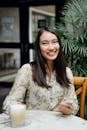Home Cheerful young Asian lady with mobile phone in casual clothes smiling and drinking latte while chilling in outdoor cafe